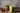 A refreshing iced matcha latte served in a mason jar with a glass straw, placed on a rustic wooden table. A small wooden bowl with matcha powder and a decorative plant in a geometric gold pot sit in the background. The text overlay reads 'Craft an Iced Ma