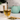 Glass of brewed Organic Healthy Colon Tea beside a wooden spoon resting on a small pile of loose herbal tea leaves, with a mint-green teapot and potted plant in the background.