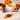 White cup of Organic Orange Cinnamon Spice tea beside a glass jar of cinnamon sticks, scattered spices, and small pumpkins with autumn leaves on a marble surface.