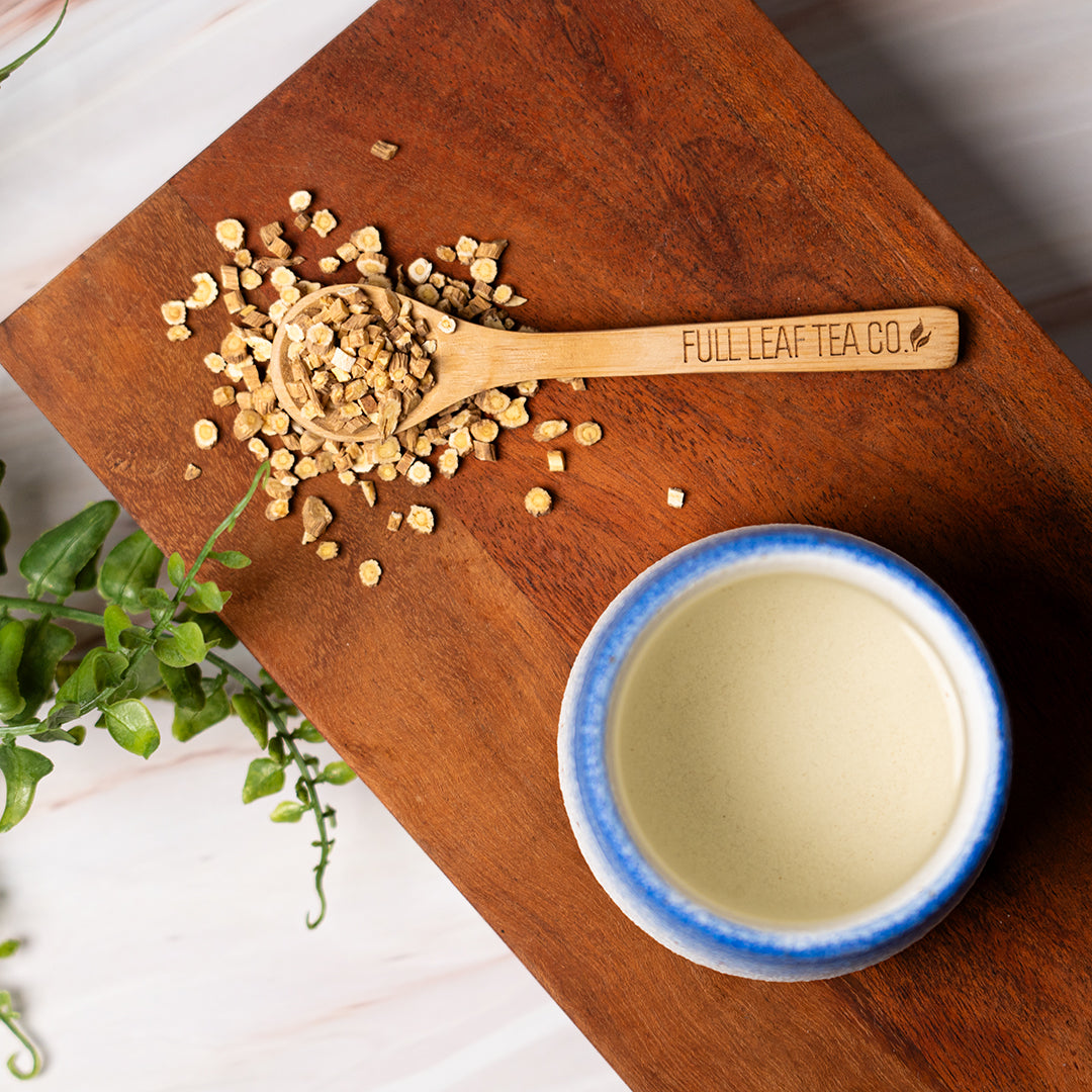 Overhead view of a wooden spoon labeled “Full Leaf Tea Co.” filled with dried astragalus root pieces on a wooden board next to a cup of tea with a blue rim and fresh green herbs.