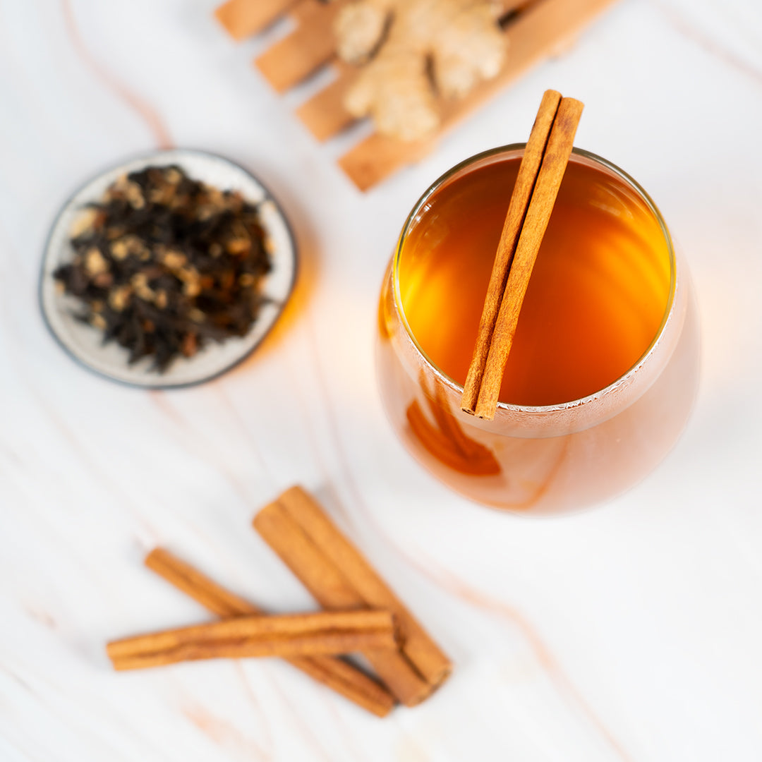 Glass of spiced tea garnished with cinnamon sticks, with loose tea leaves and ginger on a marble surface, creating a cozy and aromatic presentation.