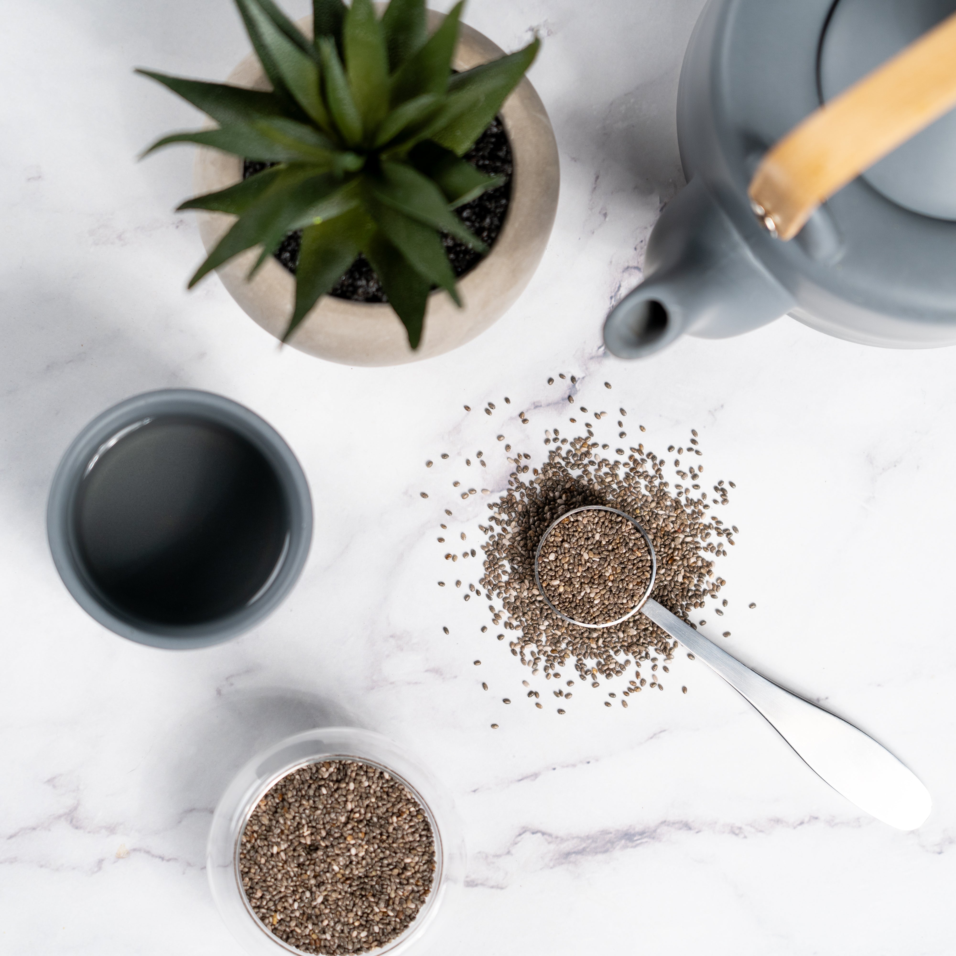 Overhead view of a metal spoon filled with chia seeds on a white marble surface, surrounded by scattered seeds, a gray teapot, a small cup, and a potted succulent plant.