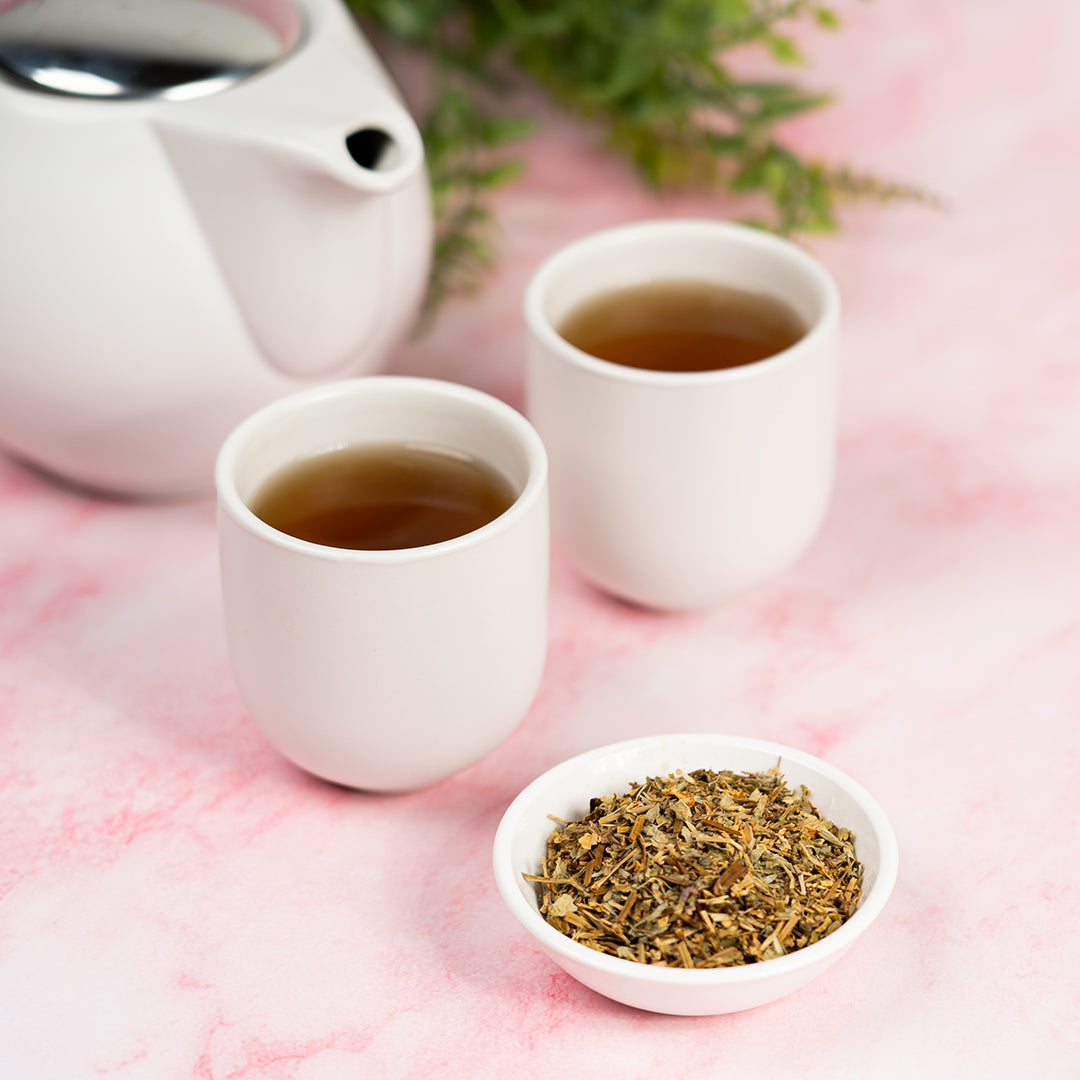 Two white cups of brewed herbal tea beside a small dish of dried chickweed leaves and a white teapot, set on a pink marble surface with greenery in the background.