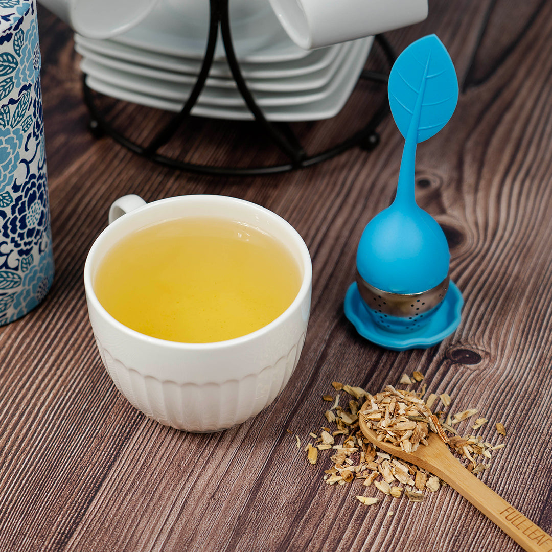 White ceramic cup of brewed chicory root tea on a wooden table beside a blue silicone tea infuser, dried chicory root pieces, and a bamboo spoon engraved with 'Full Leaf.'