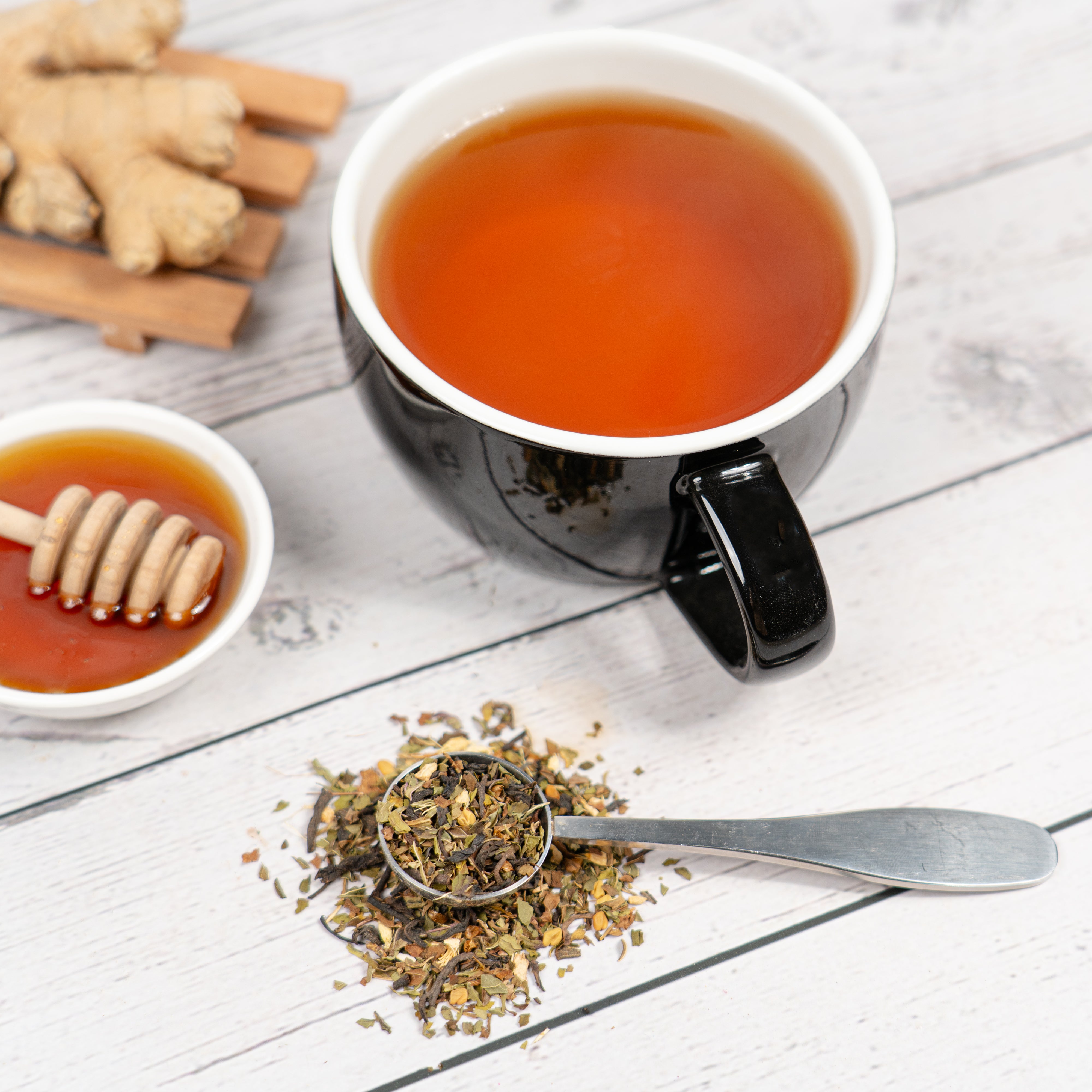 Black ceramic mug filled with hot tea on a light wooden surface, surrounded by a bowl of honey with a honey dipper, fresh ginger, and a metal teaspoon holding loose herbal tea leaves.