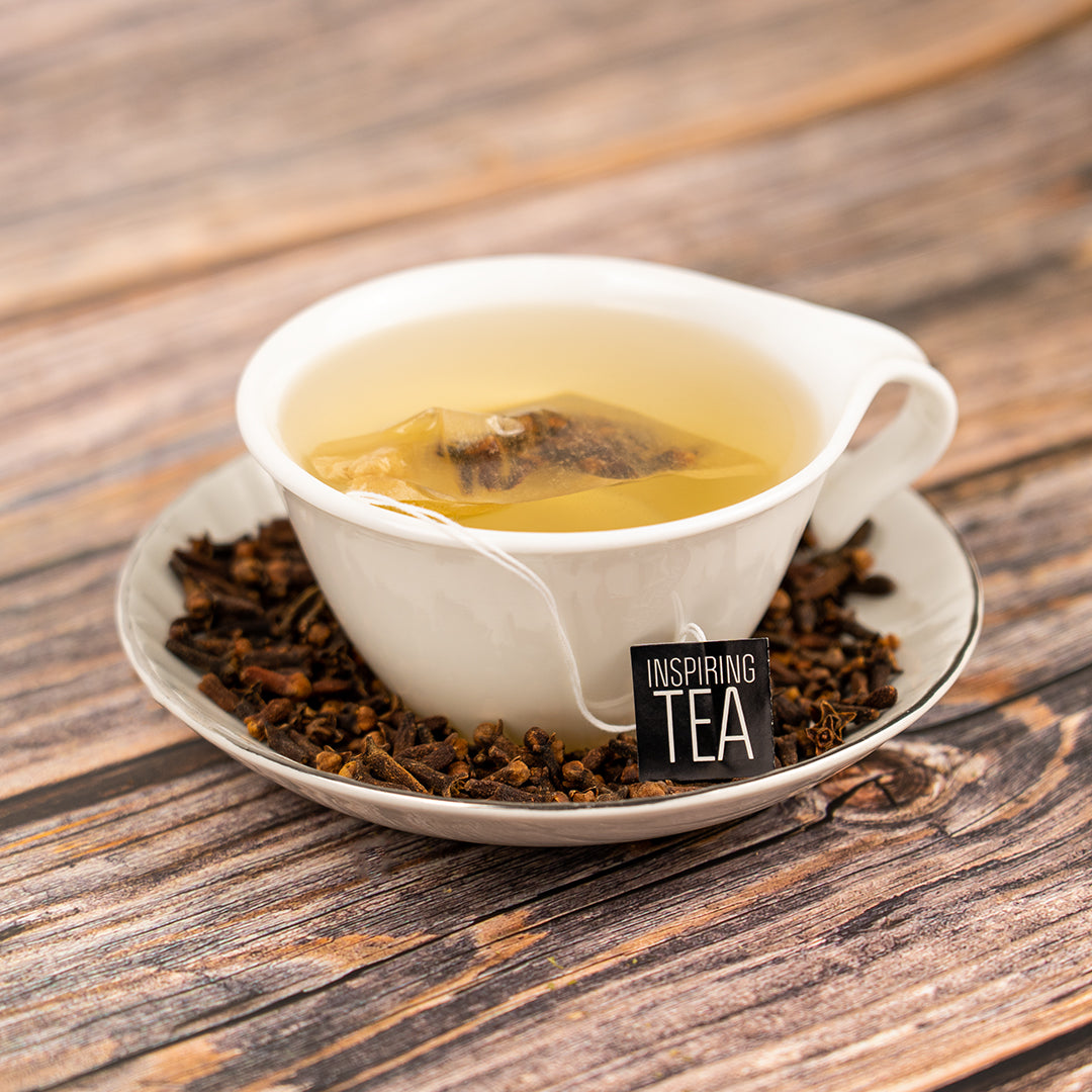 A white teacup filled with clove tea and a tea bag labeled “Inspiring Tea,” sitting on a saucer surrounded by whole cloves on a wooden surface.
