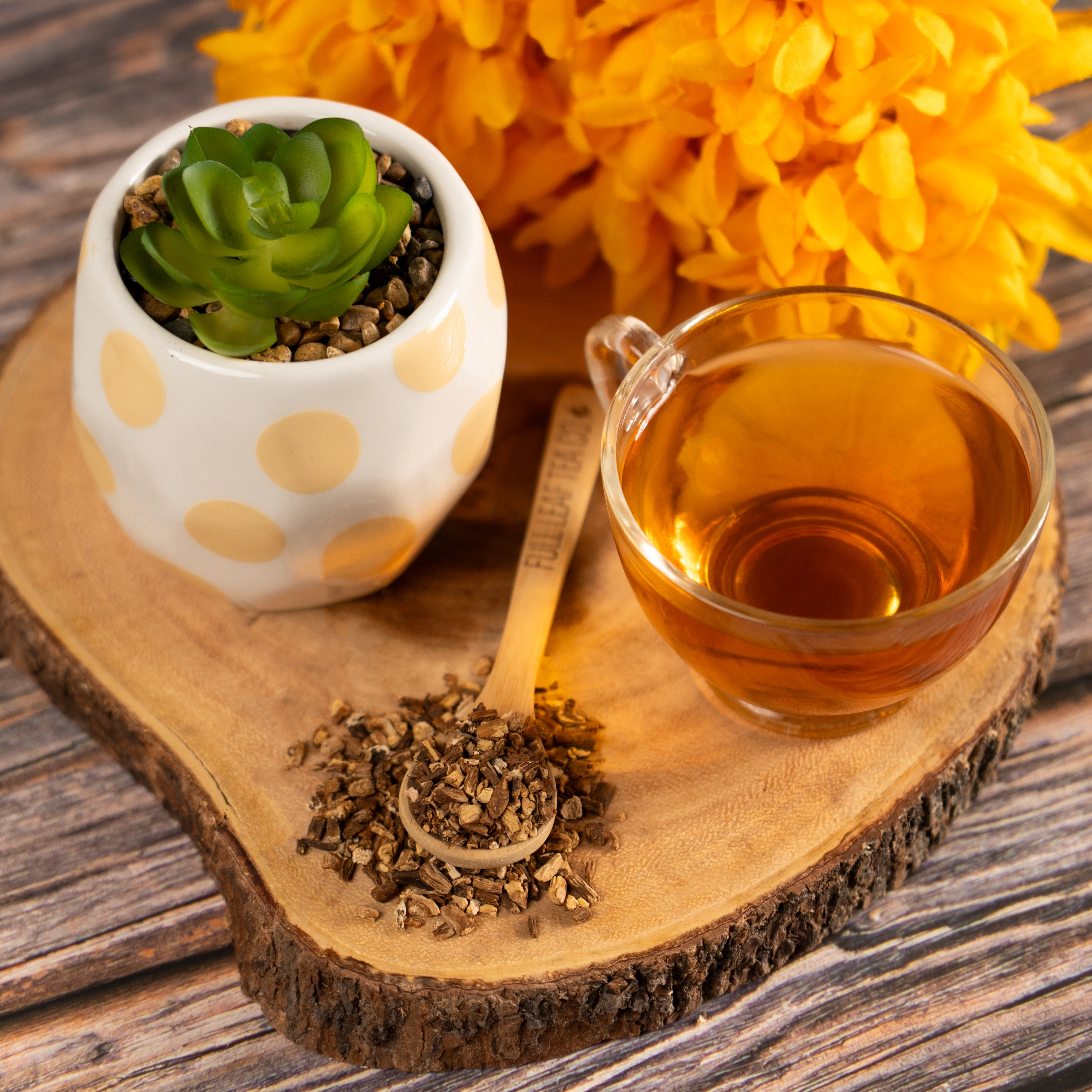 Glass cup of brewed dandelion tea on a wooden serving board beside a small scoop of dried dandelion root and a white pot with a green succulent, with bright yellow flowers in the background.
