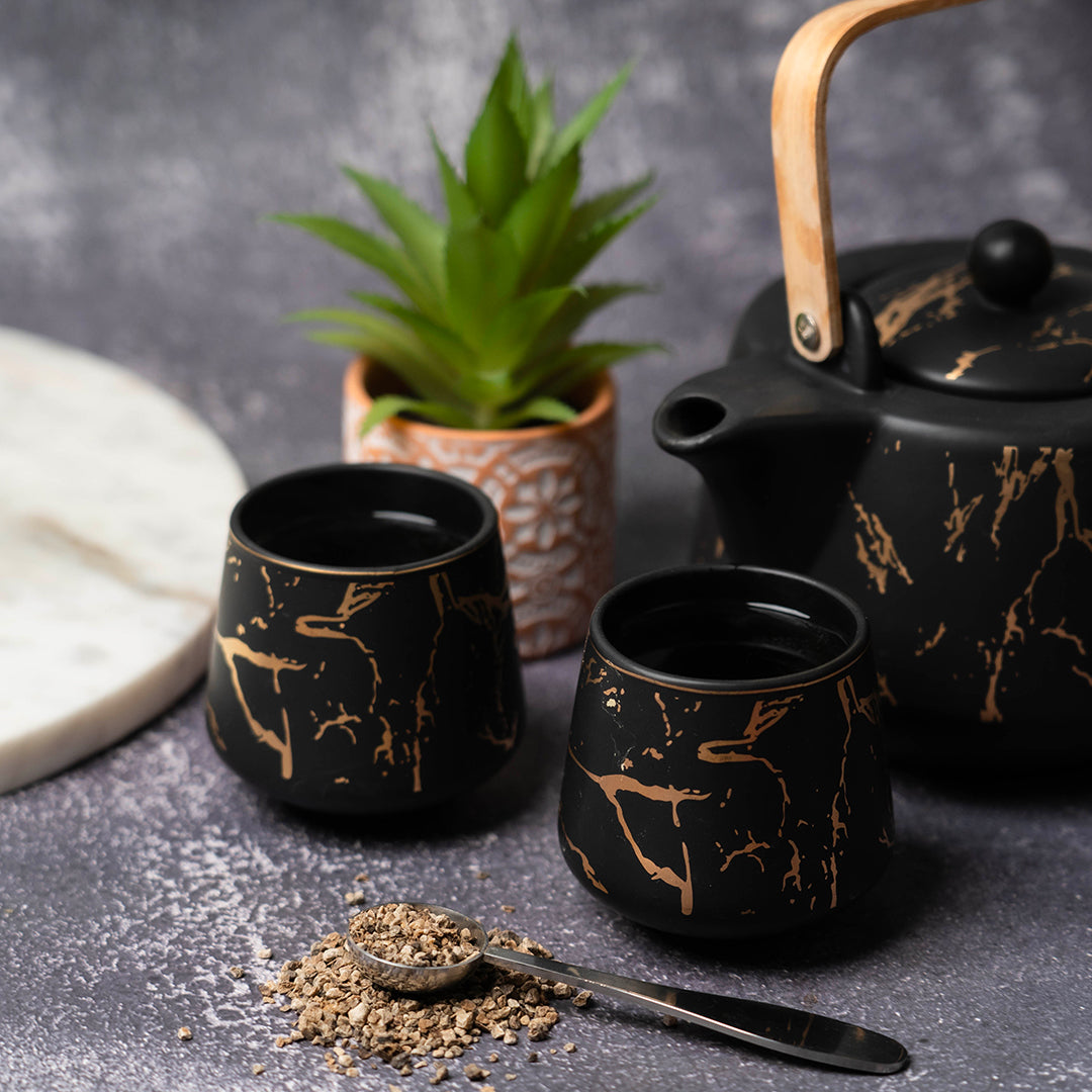A black marble-patterned teapot with a wooden handle and two matching teacups on a gray surface, with a small succulent plant and a teaspoon of devil’s claw root beside them.