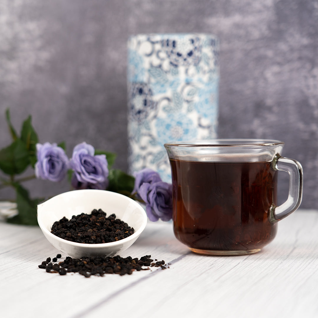 A clear mug filled with Elderberry tea with flowers in the background and a elderberries besides the mug 