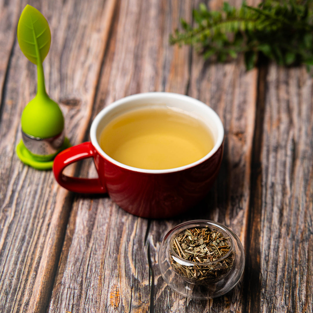 A red mug filled with freshly brewed epimedium tea on a rustic wooden surface, beside a glass bowl of dried epimedium leaves and a green leaf-shaped tea infuser.