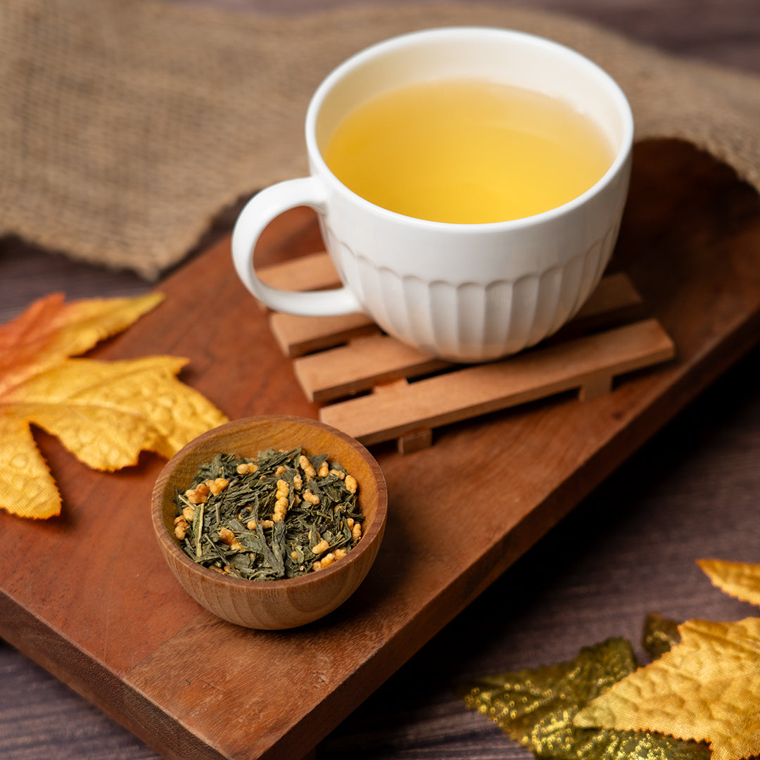 Cup of freshly brewed Organic Genmaicha tea with a small wooden bowl of loose leaf tea on a wooden tray with autumn leaves.