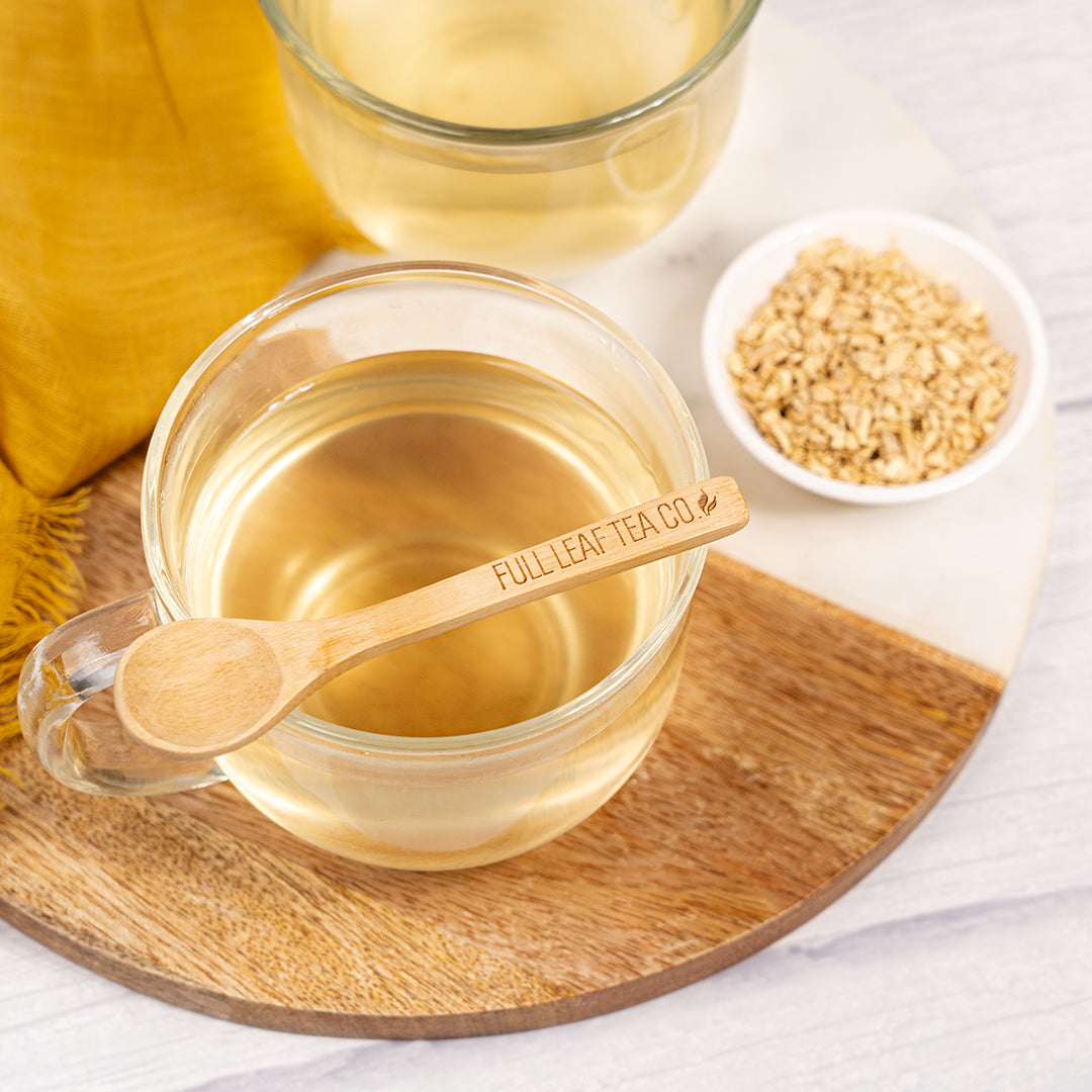 Cup of ginger tea on a wooden tray with a wooden spoon engraved ‘Full Leaf Tea Co.’ resting on top, next to a small bowl of dried ginger pieces.