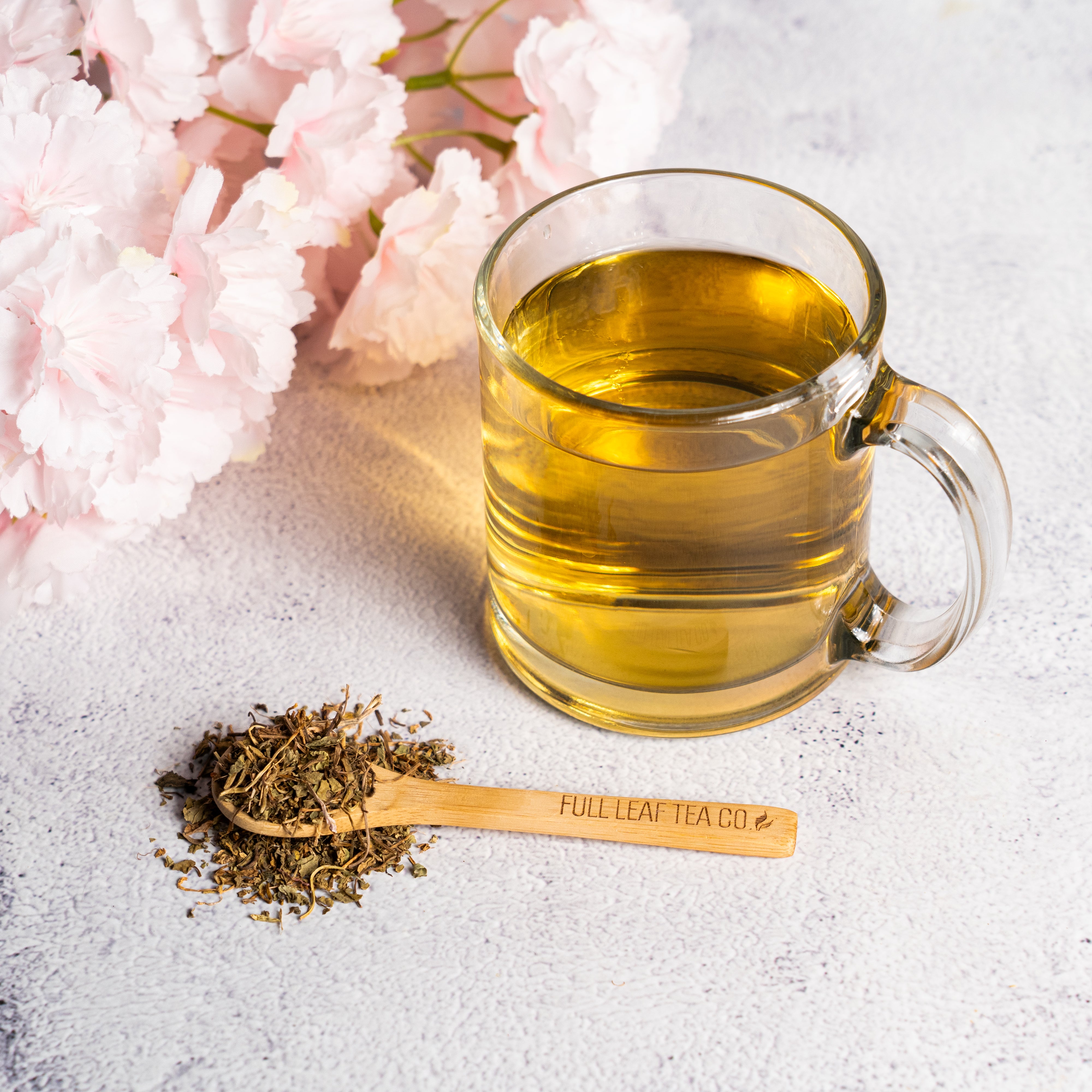Glass mug of brewed Gotu Kola tea on a light surface beside a wooden scoop labeled “Full Leaf Tea Co.” filled with loose Gotu Kola leaves, with soft pink flowers in the background.