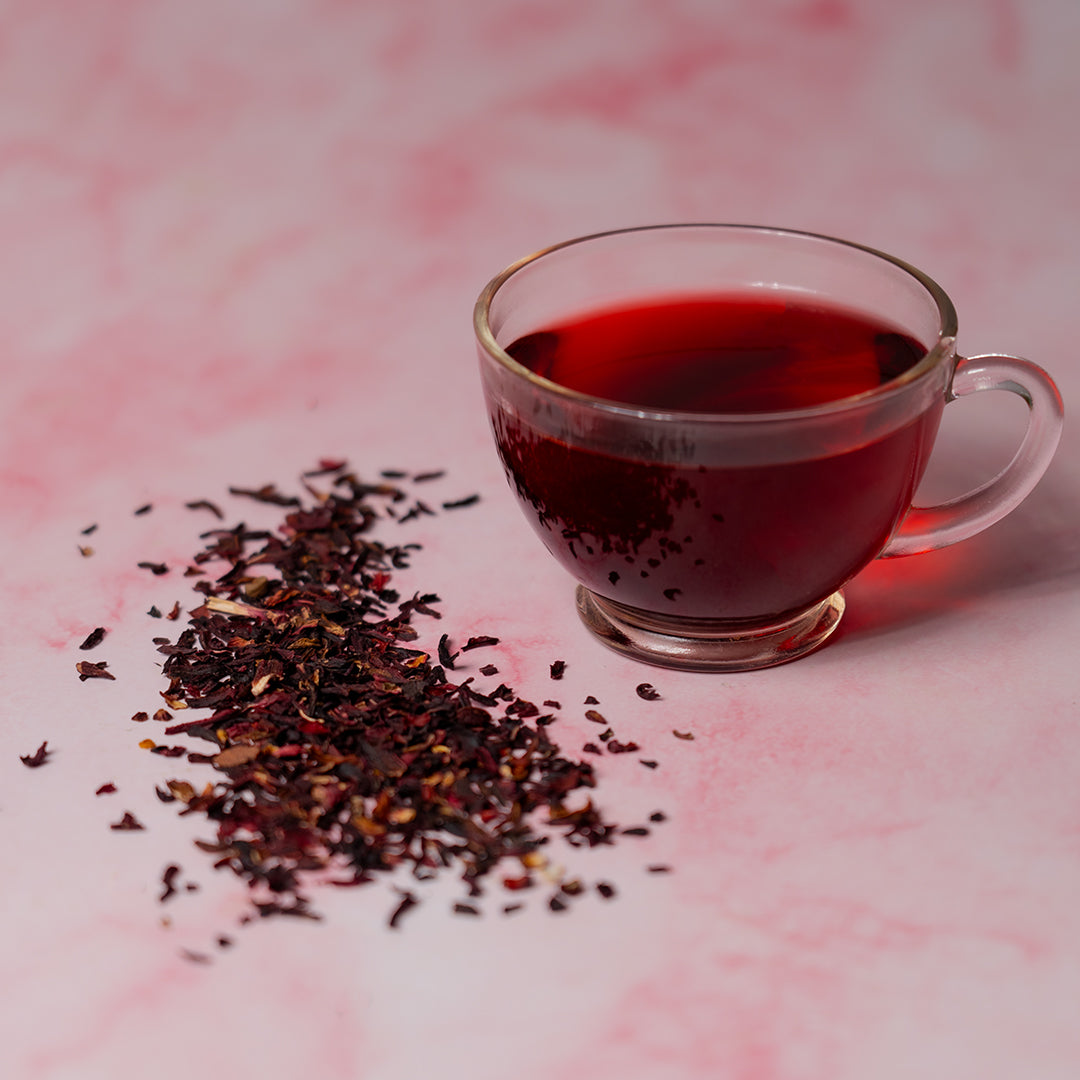 Glass cup of Organic Hibiscus Tea with deep red color beside loose dried hibiscus petals on a pink marble surface.