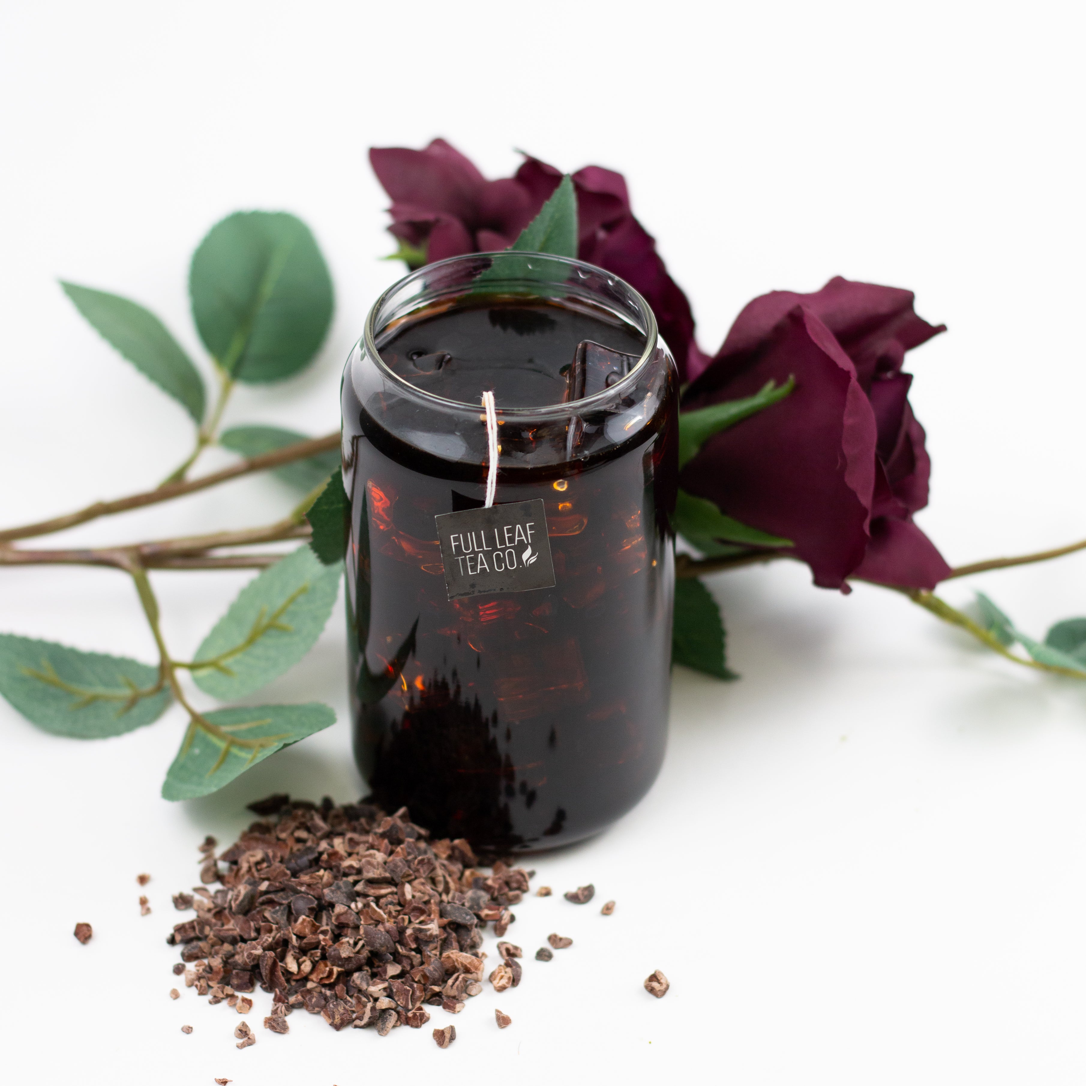 Glass jar with a tea bag string labeled ‘Full Leaf Tea Co.’ filled with tea, placed beside dried cocoa nibs  and deep purple roses on a white background.