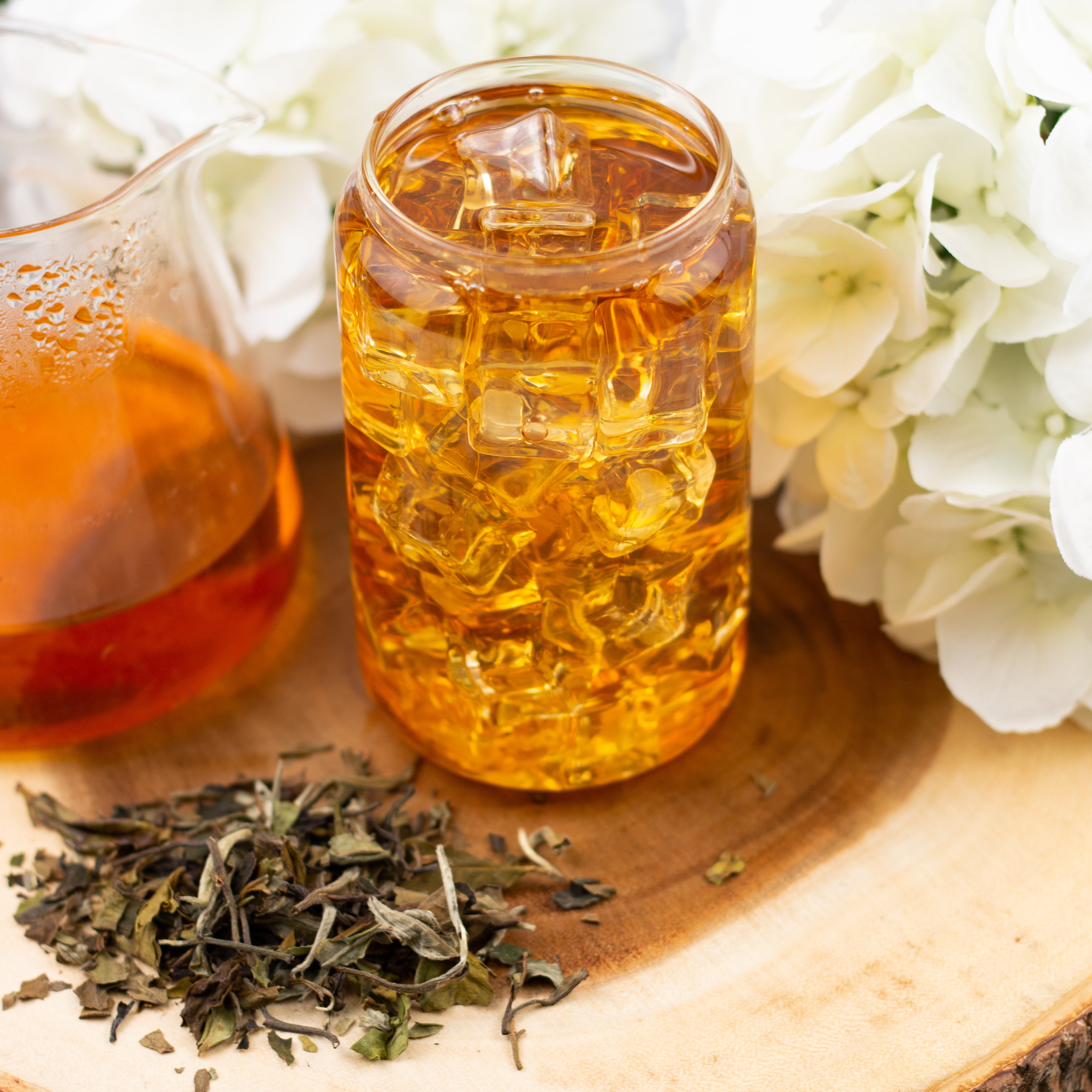 Glass of iced tea with golden amber color, surrounded by loose leaf tea and white flowers on a wooden surface.