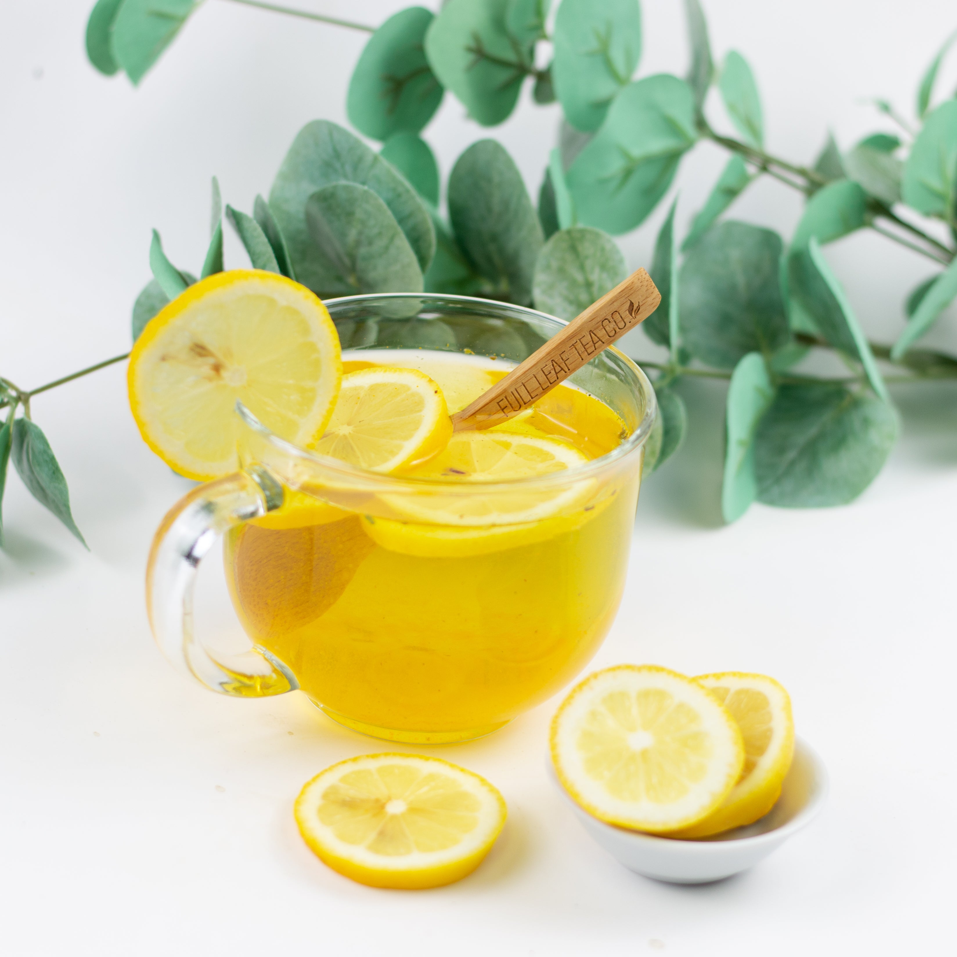 Glass mug of lemon tea with lemon slices and a cinnamon stick, surrounded by green leaves on a light background.