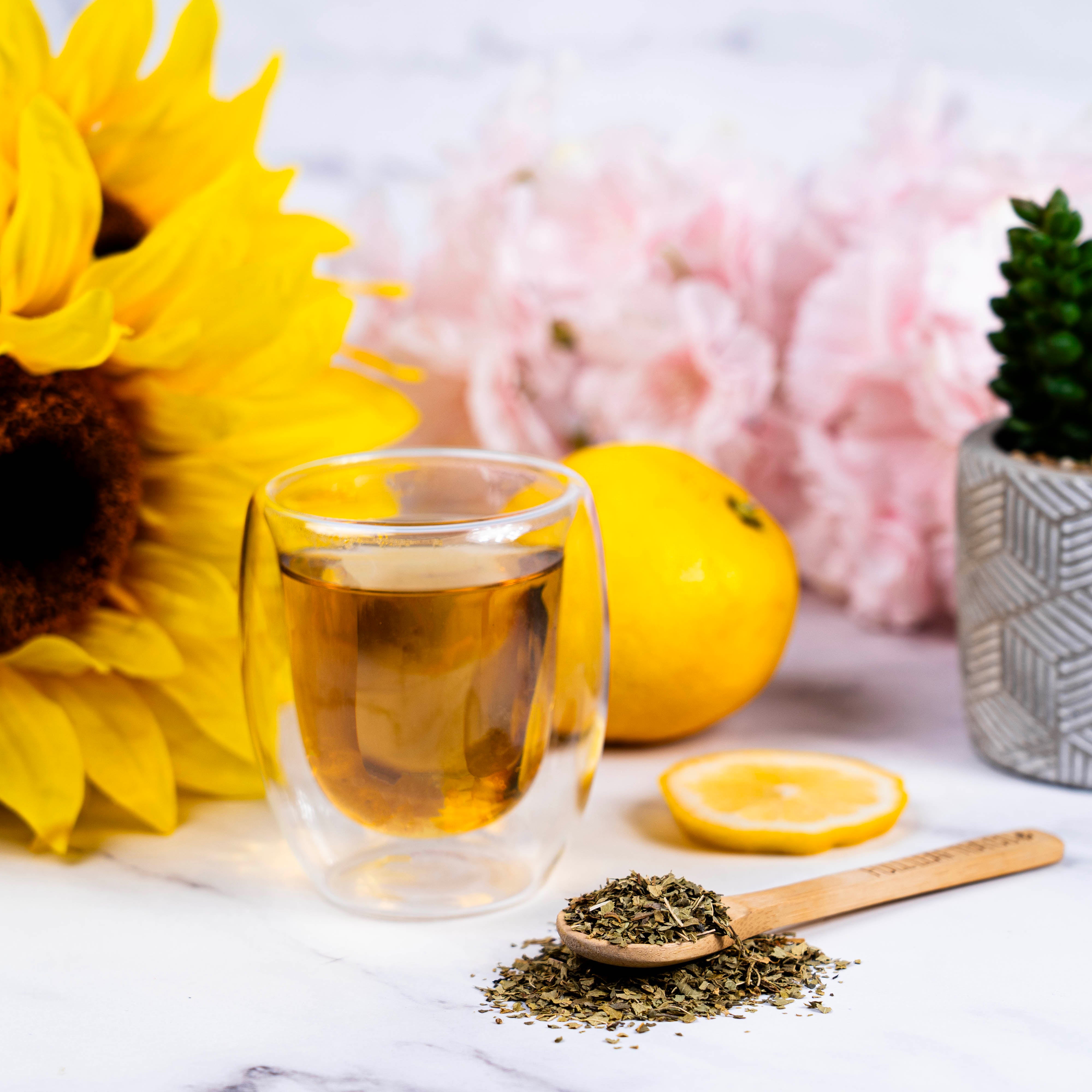 Glass cup of Organic Lemon Verbena Tea with dried tea leaves, lemon slices, and bright flowers including a sunflower on a marble surface.