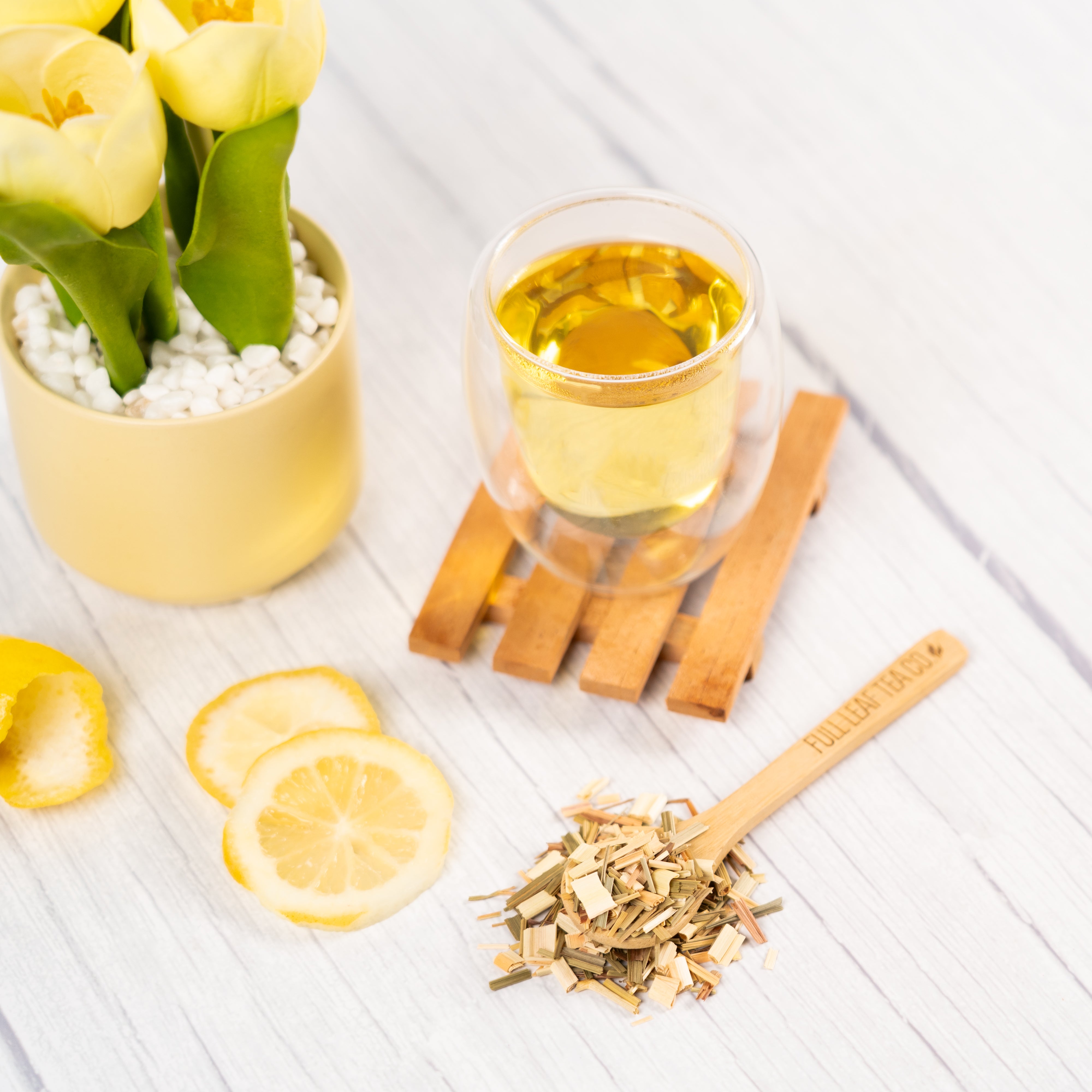 Glass cup of Organic Lemongrass Tea on a wooden coaster with dried lemongrass pieces, lemon slices, and yellow flowers on a light wooden surface.