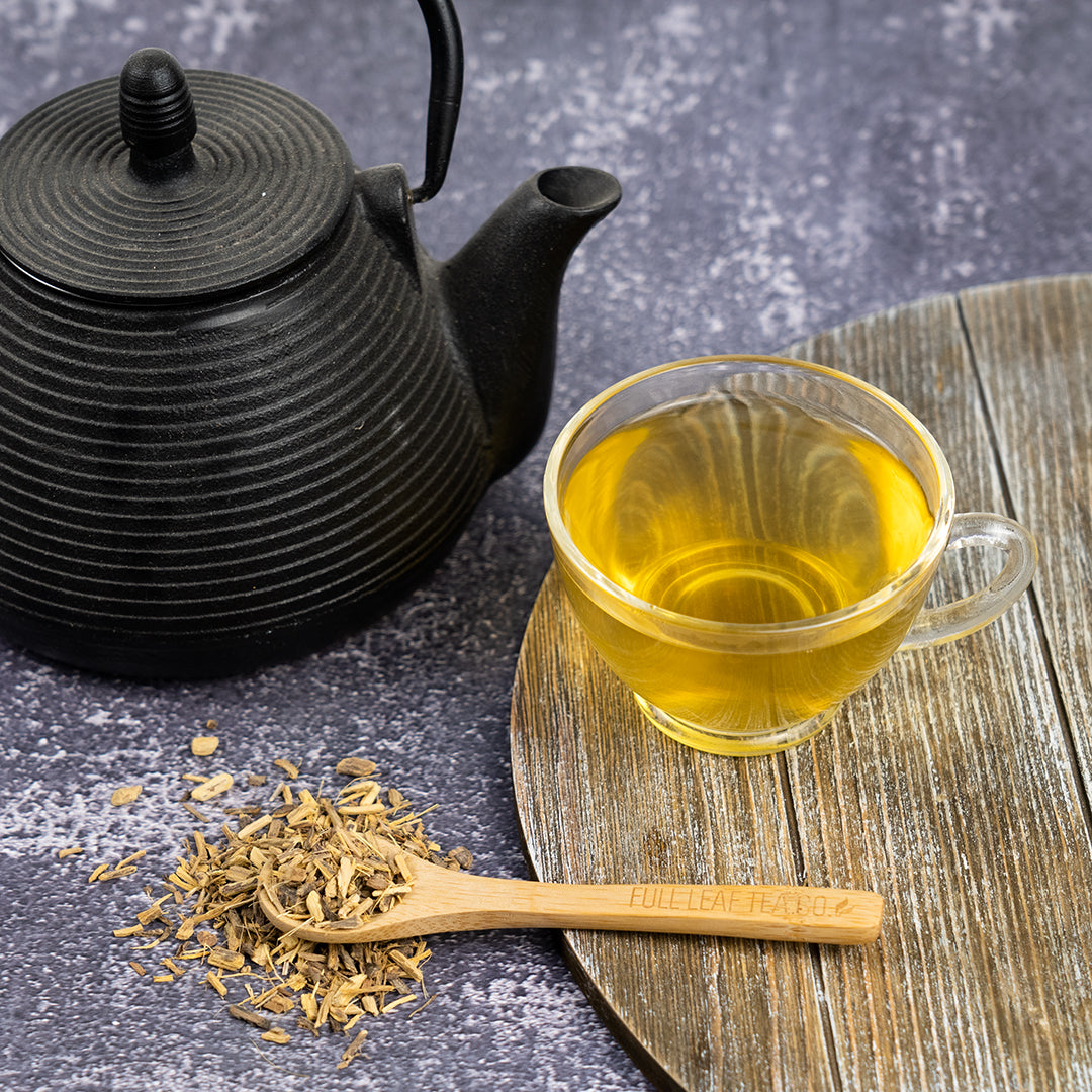 A clear mug filled with Licorice root tea on a wooden surface surrounded by a black tea pot and licorice root