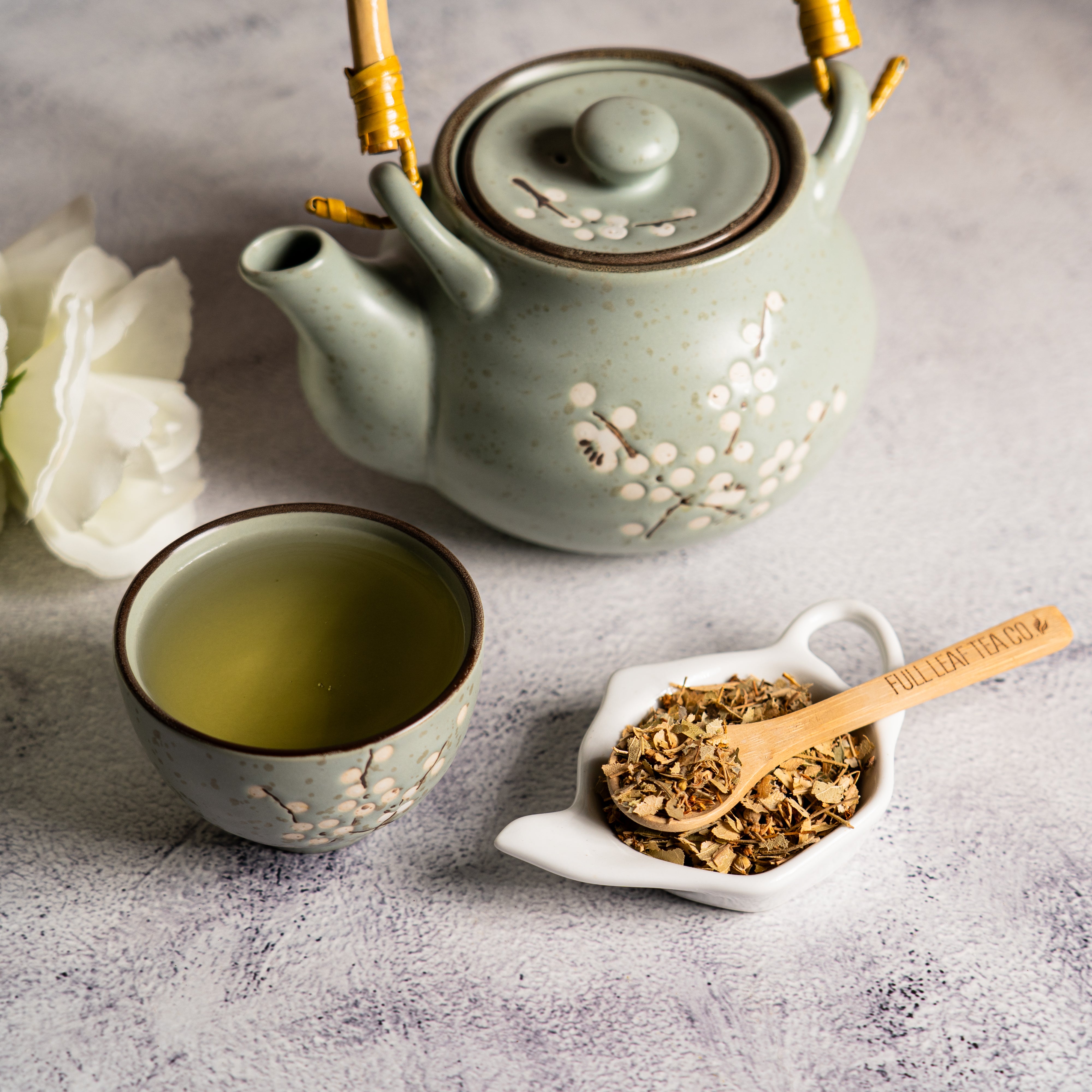 Cup of brewed Organic Linden Leaf tea with dried linden leaves on a small white dish and a ceramic teapot with floral design on a textured surface.