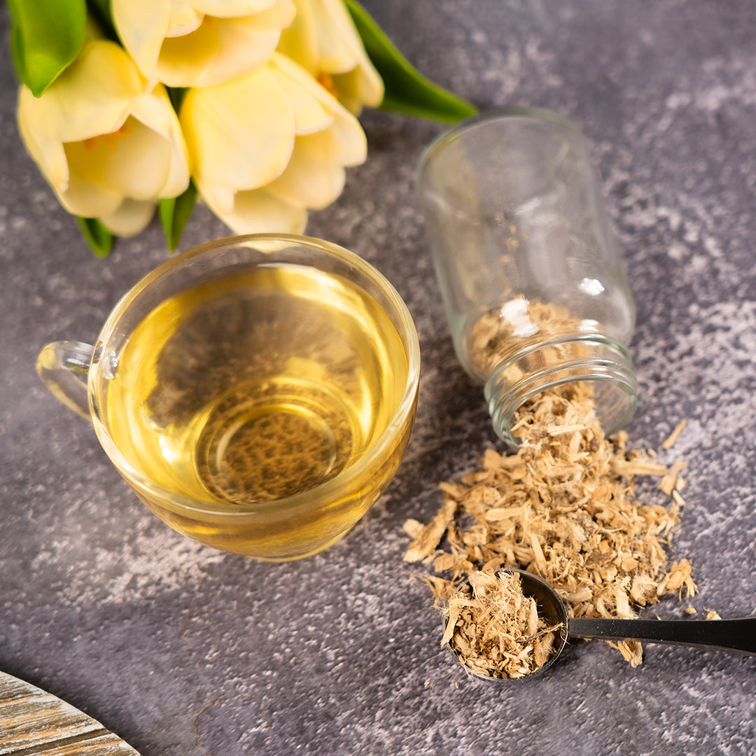 Cup of brewed marshmallow root tea beside a glass jar spilling dried marshmallow root pieces and a measuring spoon on a gray textured surface with yellow tulips in the background.