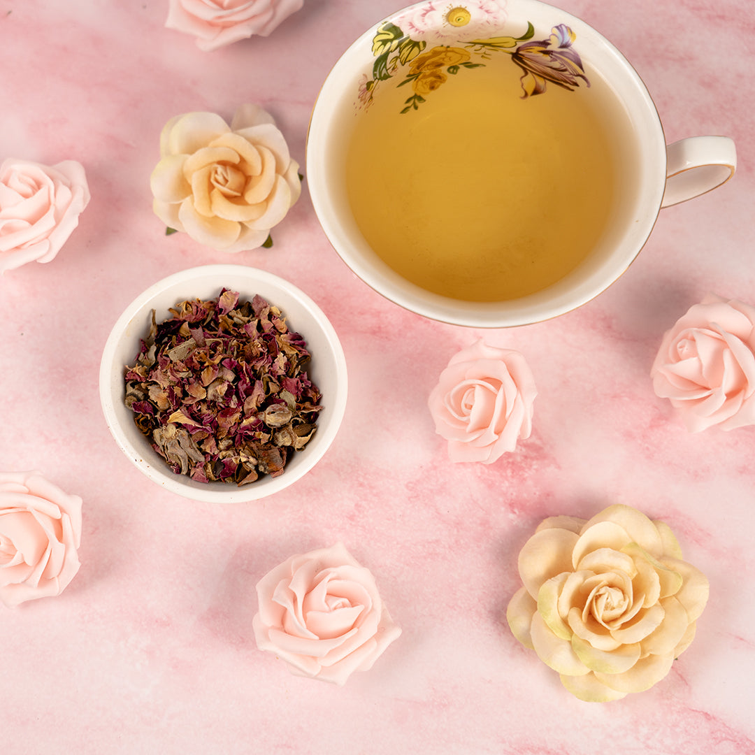 A cup of Rose Petal Tea in a white mug with a bowl of rose petals beside it and the surface is marble with rose petals on it. 