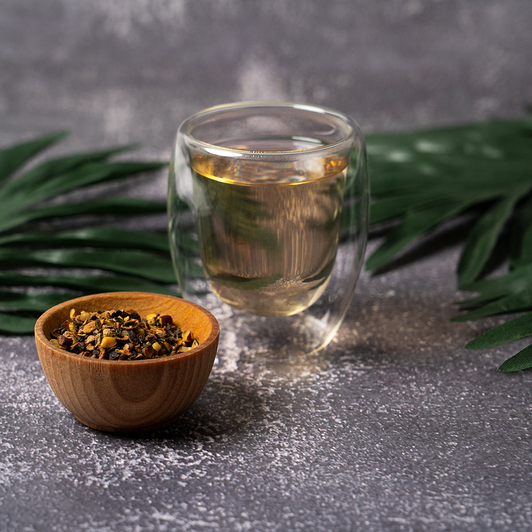 Glass cup of Organic Saw Palmetto tea beside a wooden bowl of dried Saw Palmetto ingredients, with green palm leaves in the background.