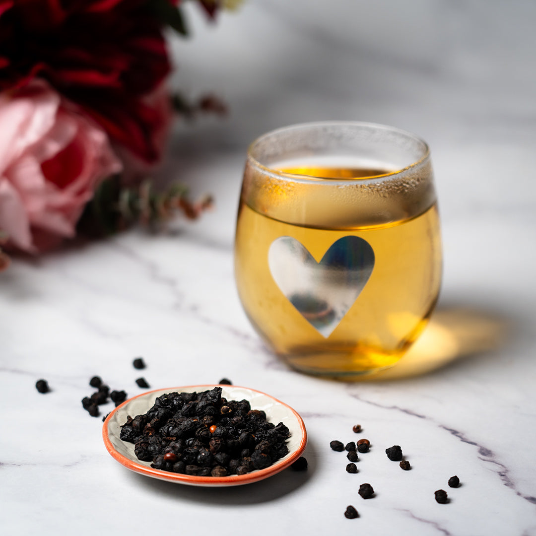 Glass cup of Organic Schisandra Berry tea with a heart design, beside a small dish of dried black Schisandra berries on a marble surface.