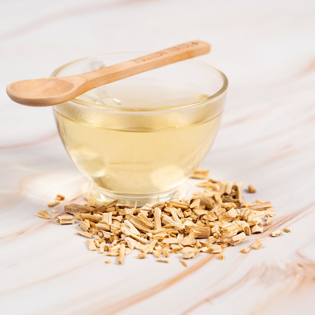 Cup of Organic Shatavari Tea with light golden color, surrounded by dried shatavari root pieces and a wooden tea spoon.