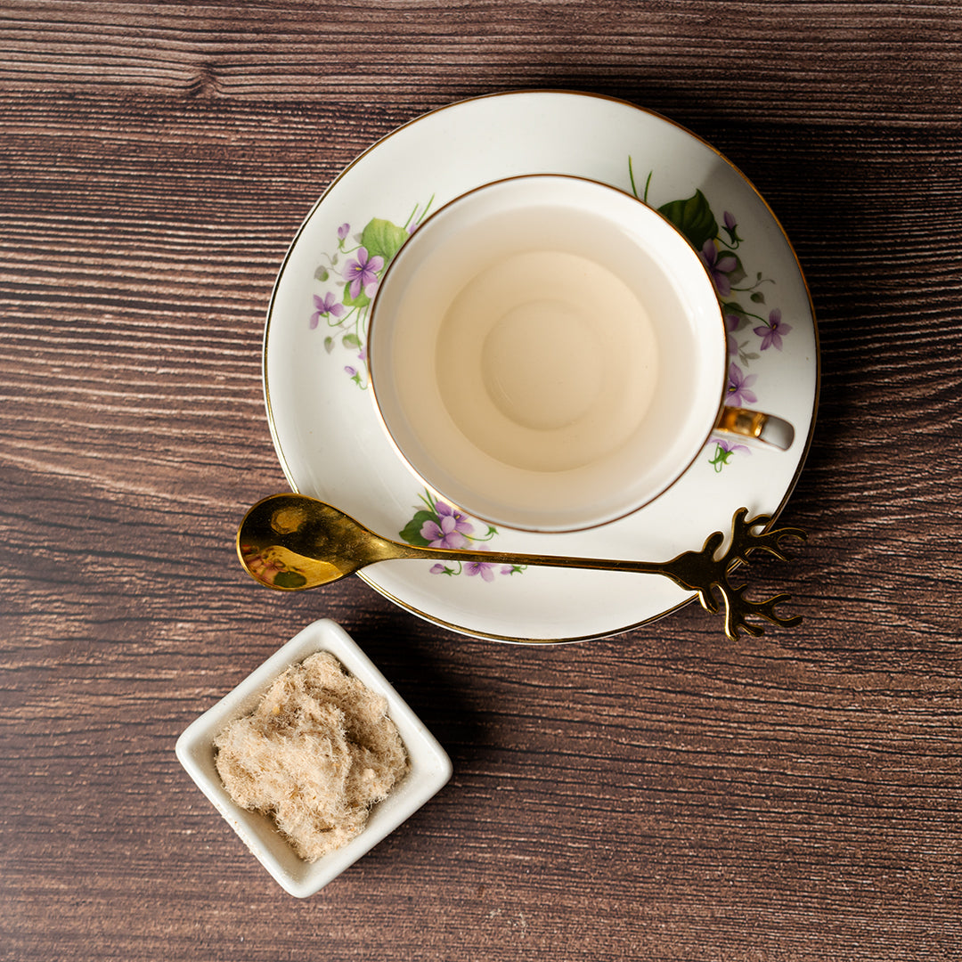 Top-down view of a cup of Slippery Elm tea in a floral teacup with gold accents on a matching saucer, beside a gold spoon and a small white bowl filled with powdered Slippery Elm bark on a wooden surface.