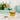 Glass of spearmint tea with a small bowl of spearmint leaves on a marble surface, with a blurred background of kitchen utensils and plants.