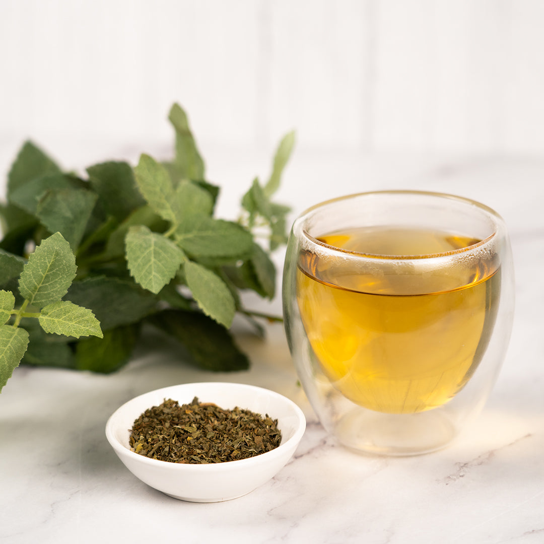 Glass cup of spearmint tea with light golden color on a white surface beside a small bowl of dried spearmint leaves and a bunch of fresh green spearmint leaves in the background.