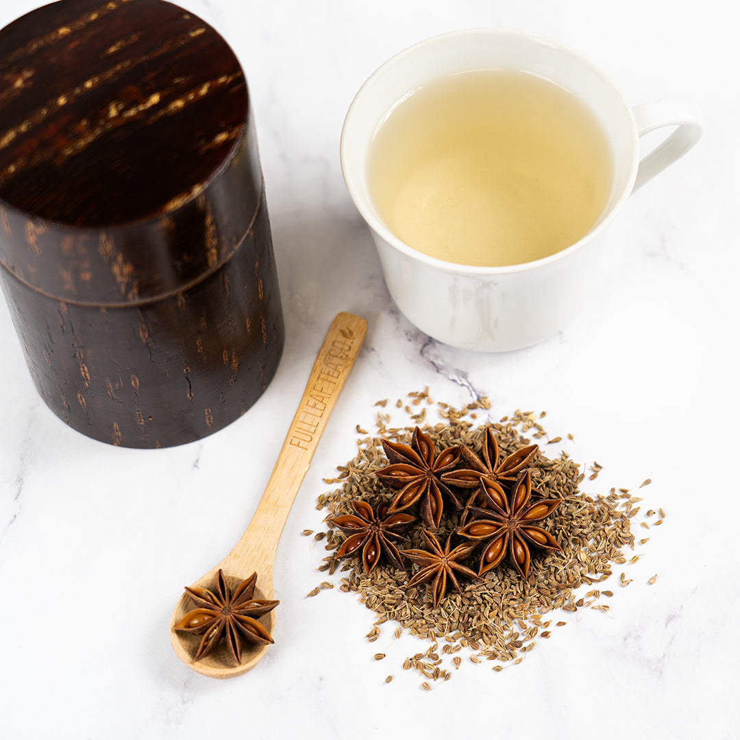 A white cup of brewed star anise tea beside a dark wooden tea canister and a wooden spoon labeled “Full Leaf Tea Co,” surrounded by dried star anise pods and seeds on a white marble surface.