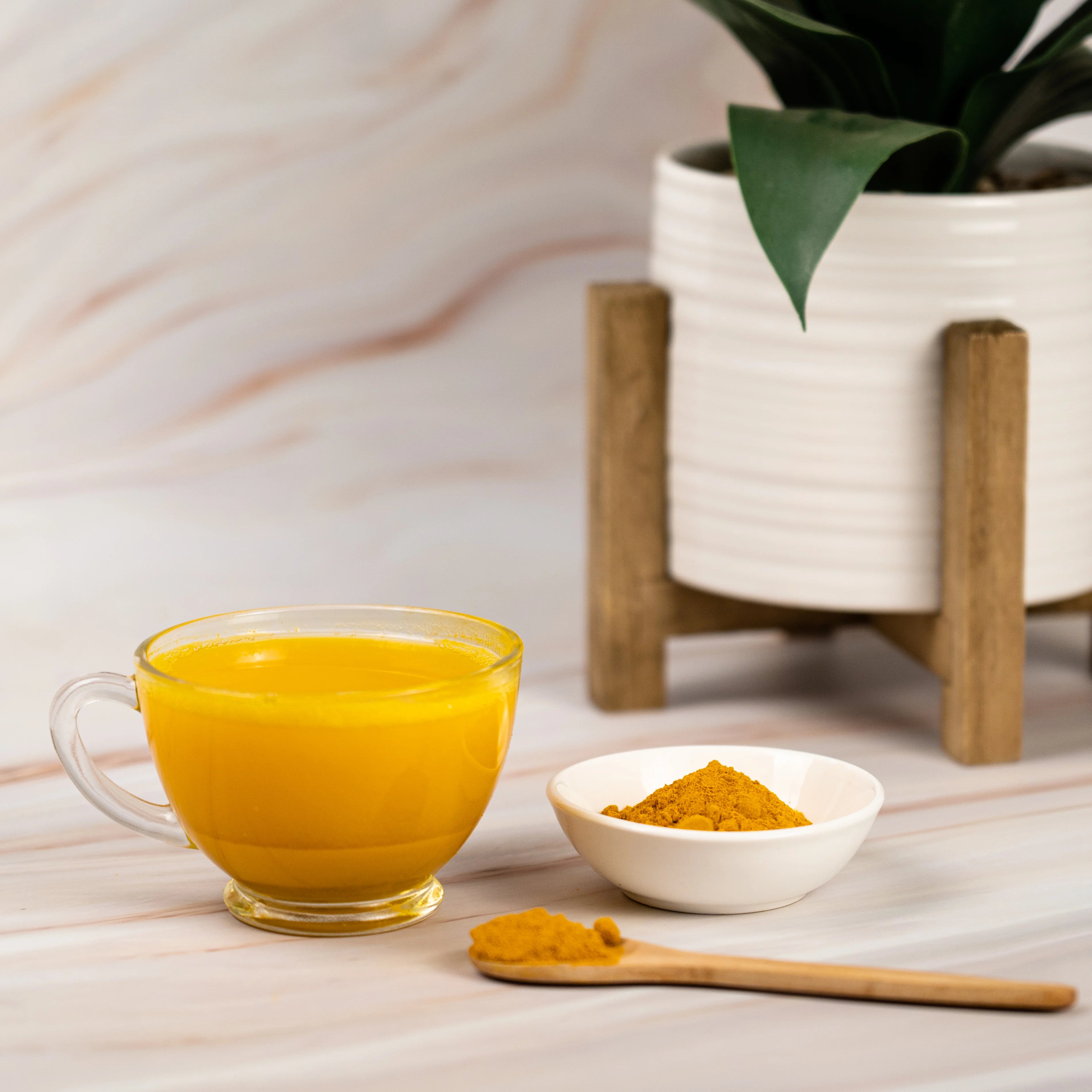 A glass cup of turmeric tea beside a small white bowl and wooden spoon filled with turmeric powder, with a potted plant in the background on a light marble surface.