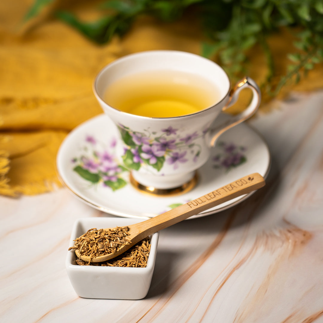 Delicate floral teacup filled with golden valerian root tea on a matching saucer, beside a wooden spoon labeled ‘Full Leaf Tea Co.’ resting on a small white dish of dried valerian root, with a soft yellow cloth and green leaves in the background.