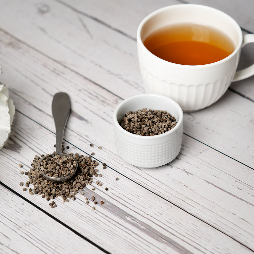 White teacup filled with vitex berry tea beside a small white bowl and metal spoon filled with dried vitex berries on a light wooden surface.