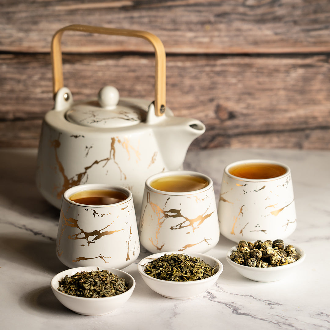 Elegant ceramic teapot with matching cups filled with tea, surrounded by bowls of loose leaf green tea on a marble surface.