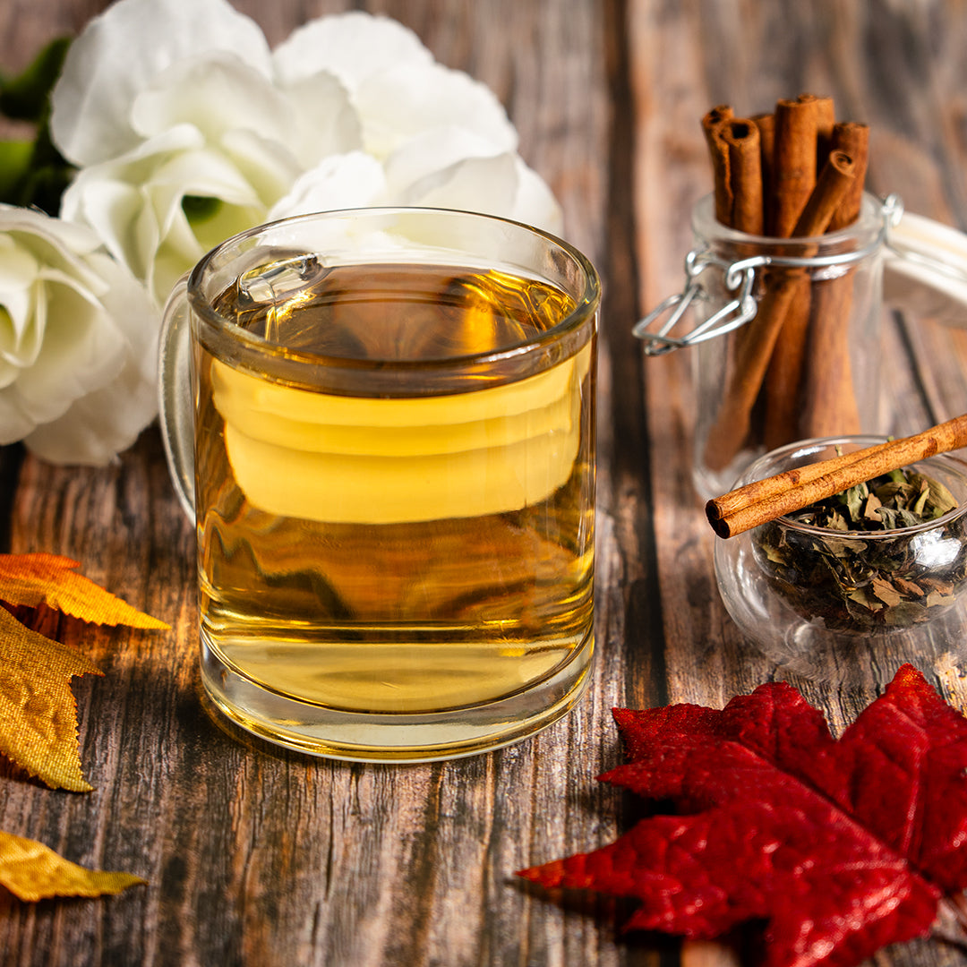 Glass mug of golden Organic White Chai with cinnamon sticks, loose leaf tea, and autumn leaves on a rustic wooden table.