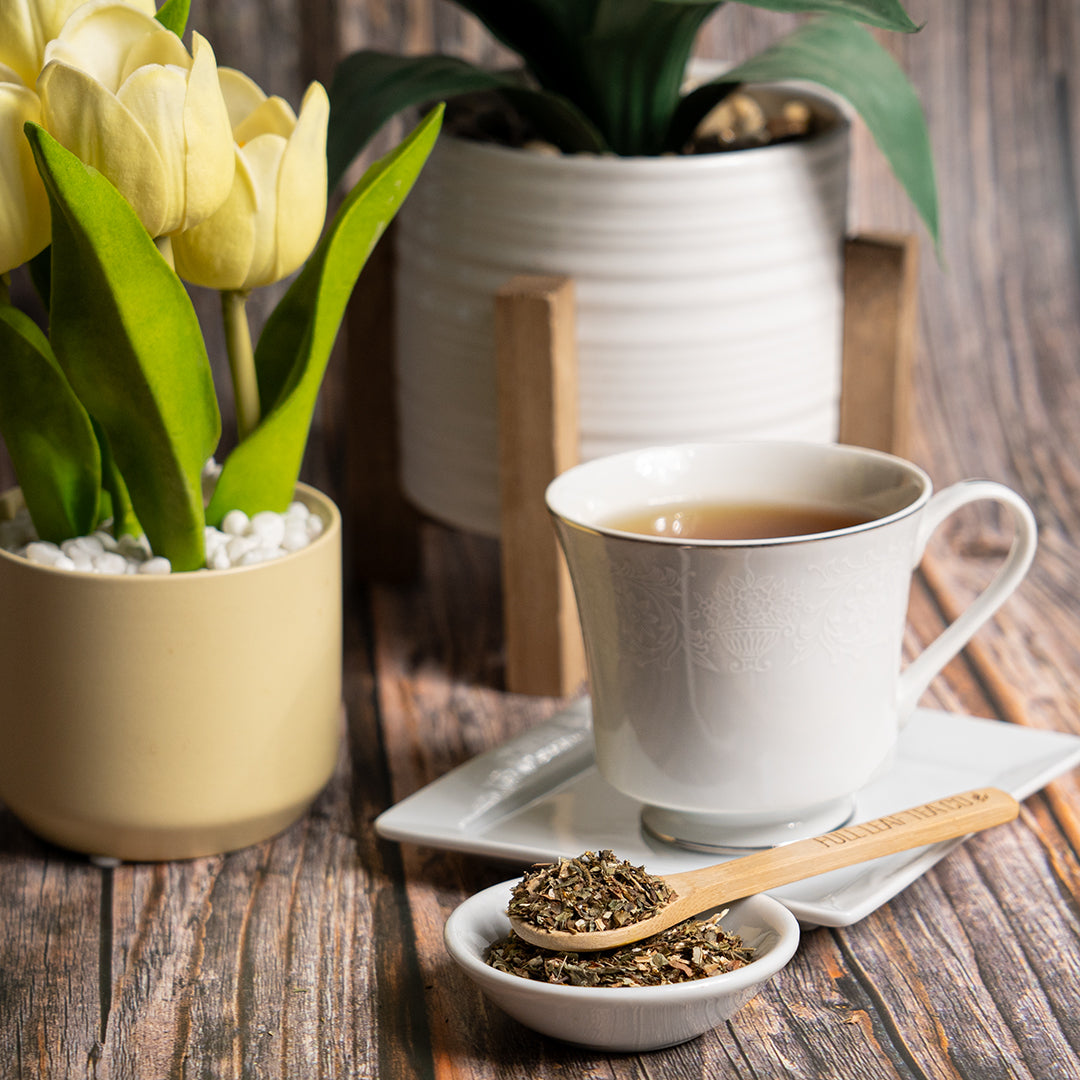 White teacup filled with wild lettuce tea on a square saucer beside a wooden spoon labeled ‘Full Leaf Tea Co.’ holding dried wild lettuce leaves, with potted plants in the background on a rustic wooden surface.