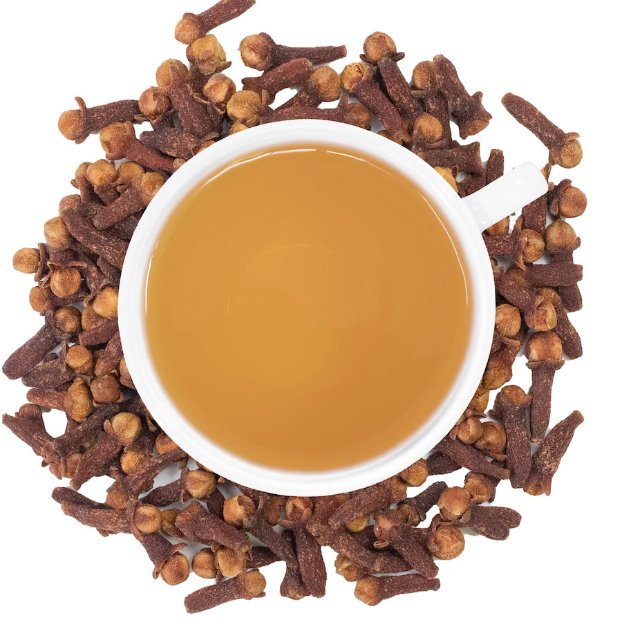 A white mug filled with clove tea surrounded by cloves on a white surface. 