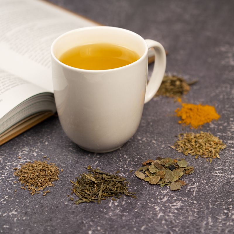 White mug of Organic Healthy Vision Tea beside an open book, surrounded by small piles of loose tea ingredients including herbs, leaves, and turmeric on a gray textured surface.