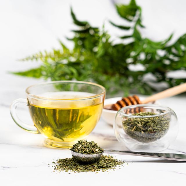 Glass cup of brewed nettle tea with loose dried nettle leaves, a metal teaspoon, and a honey dipper on a marble surface with green ferns in the background.