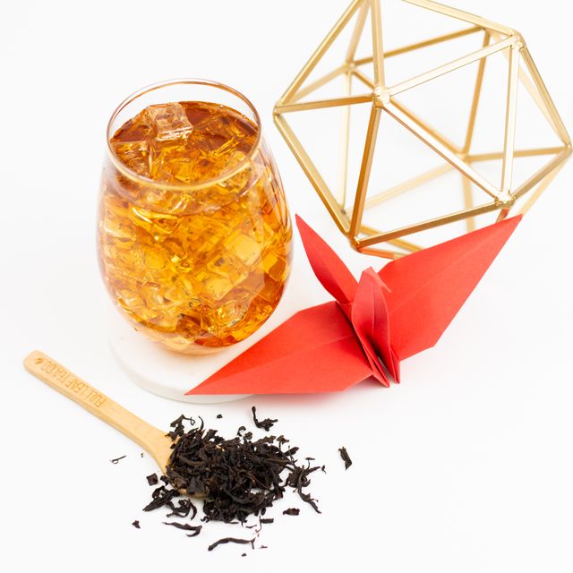 Glass of iced oolong tea with loose leaf tea and a wooden scoop beside a red origami crane and geometric gold decor on a white background.