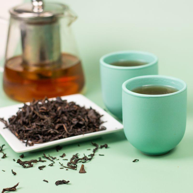 Two teal cups of brewed oolong tea beside a glass teapot and loose leaf oolong tea on a white plate against a light green background.