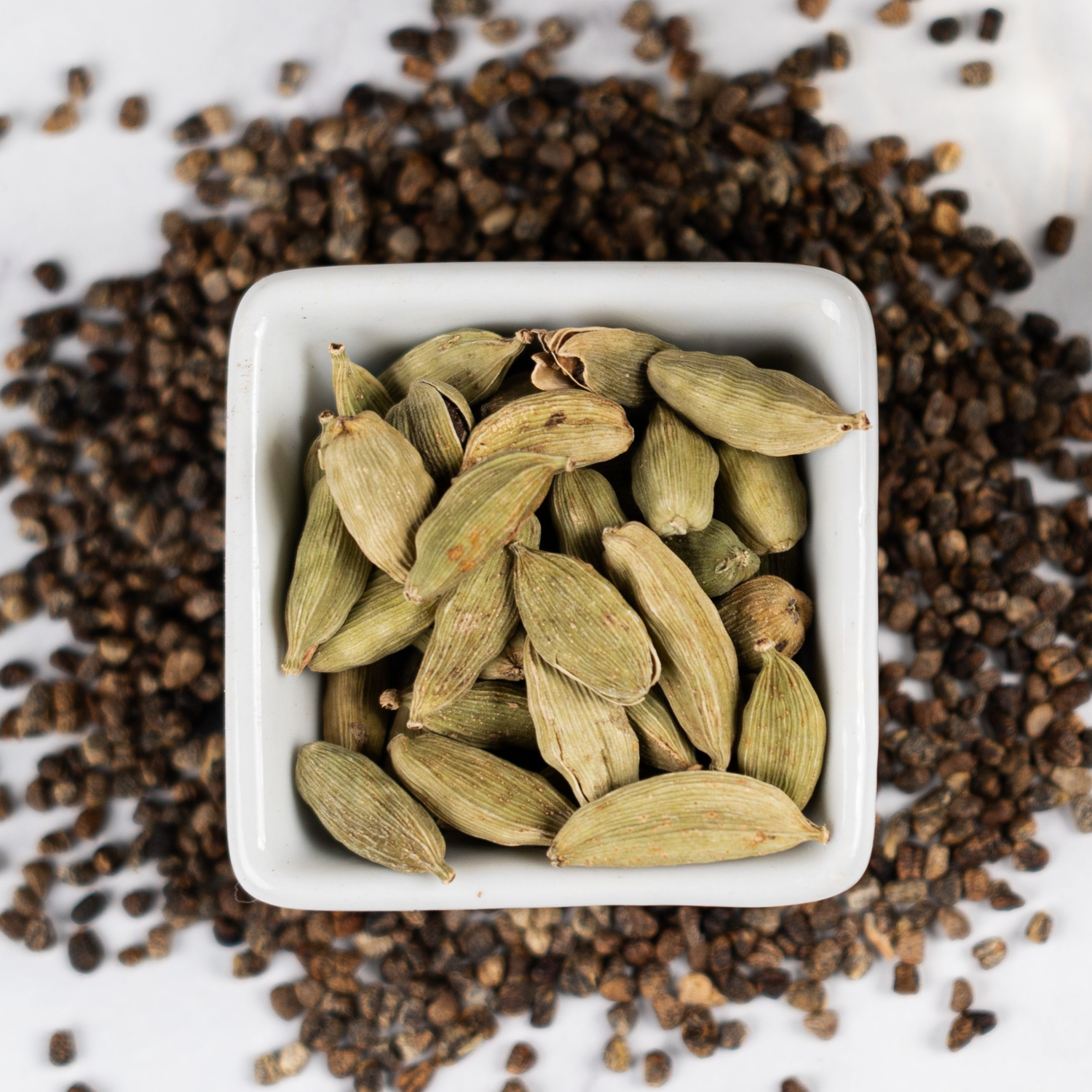 Overhead view of a bowl filled with green cardamom pods surrounded by loose black cardamom seeds on a white surface.