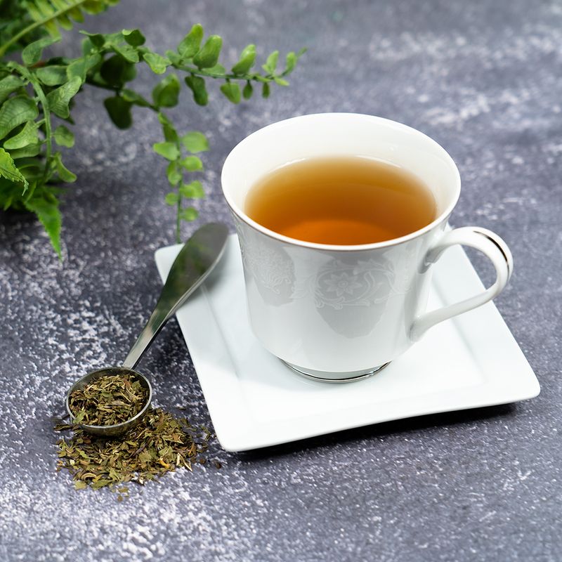 Cup of peppermint tea on a white saucer with a spoonful of dried peppermint leaves beside it, set on a gray textured surface with green foliage in the background.