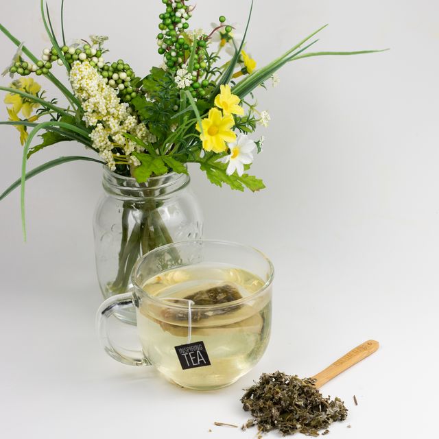 Glass cup of Organic Raspberry Leaf tea with a tea bag and loose leaves beside it, placed in front of a jar of fresh yellow and white flowers on a white background.