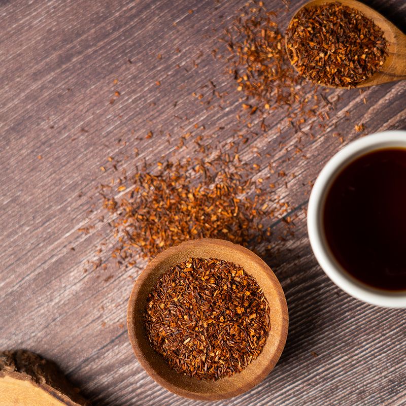 Loose rooibos tea leaves in a wooden bowl and spoon beside a cup of brewed rooibos tea on a rustic wooden surface.