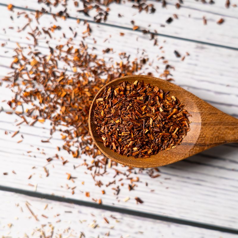 Close-up of loose rooibos tea leaves in a wooden spoon on a white wooden background.